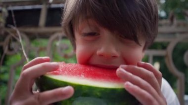 Genç çocuk bir dilim karpuzun tadını çıkarıyor, iki eliyle büyük kabuğu tutuyor. Neşeli ifadesi, doğal bir ortamda ferahlatıcı bir yaz atıştırmasının zevkini yansıtıyor.