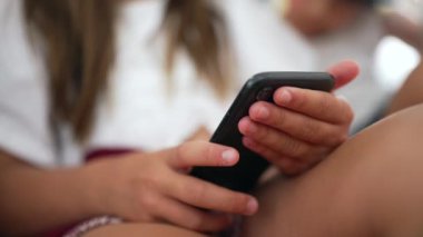 Akıllı telefonu tutan genç bir kızın hassas tutuş şekline odaklandığı yakın plan. Çocukların teknolojiyle gelişigüzel iç mekanlarda etkileşimi