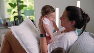 Anne, bebeğini kucaklarken sevgiyle öpüyor. Huzurlu, rahat bir ev ortamında samimi bir sıcaklık ve şefkat anı yaşıyor.