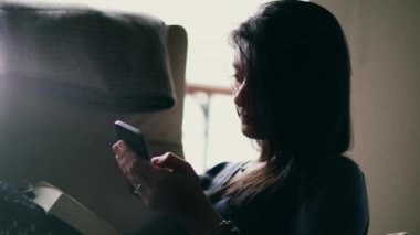 Kanepede oturan bir kadın, akıllı telefonunda gezinen, düşüncelere dalmış. Yakındaki kitap, teknolojiyle sakin ve düşünceli bir etkileşimin atmosferine katkı sağlıyor.