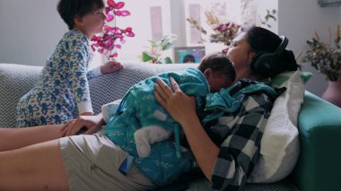 Anne, kulaklıklarla bir kanepede dinleniyor, uyuyan bir bebeği göğsünde sallarken, küçük bir çocuk yakınlarda uzanıyor, evde huzurlu ve samimi bir aile anı yaratıyor.