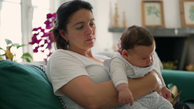 Bebeğini kollarında tutan odaklanmış bir anne, canlı iç mekan bitkileriyle çevrili bir koltukta oturuyor, sıcaklık ve bağlantıyla dolu huzurlu bir aile sahnesi yaratıyor.