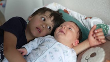 Ağabeyi yeni doğan kardeşinin yanında yatıyor, bebeğe şefkatle bakıyor, yumuşak battaniyeli bir yatağa sarılıyor, ev ortamında, aile ortamını besliyor.