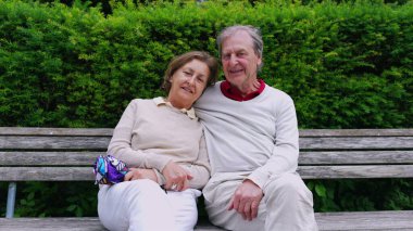 Yaşlı çift parkta bir bankta oturuyor, birbirlerine yaslanırken mutlu bir şekilde gülümsüyor, sıcak ve şefkatli bir anı yakalıyorlar.