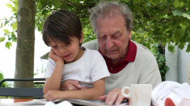 Dışarıda birlikte kitap okuyan yaşlı bir adam ve genç bir çocuk, bir ağacın gölgesinde öğrenme ve kaynaşma anını paylaştıklarında çocuk şaşkın bir ifade sergiliyor.