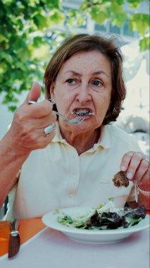 Yaşlı kadın sağlıklı bir salatanın tadını çıkarıyor. Elinde çatal dolusu yeşillik ve bir parça ekmek var. Doğal ışık ve yeşillikle çevrili.