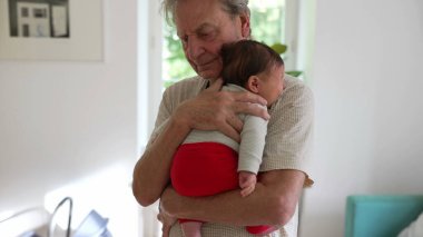Büyükbaba ağlayan küçük torununu nazikçe sakinleştiriyor, kucağında bebeği kucağında tutarken çocuk ağlıyor ve şikayet ediyor, bir yuva ortamında hassas bir rahatlık ve bakım anını vurguluyor.