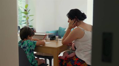 Bir anne, çocuğunu evde eğitim seansı sırasında masada işine konsantre olurken, kucağında bir bebek tutarken, sevgi dolu, eğitici bir aile anını yakalar.