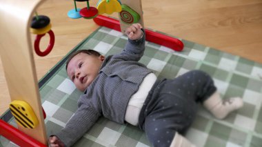 Bebek bir aktivite minderinde yatıyor, renkli asılı oyuncaklarla oynamak için uzanıyor. Oyuncu ortamı, bebeğin bakım evindeki merakını ve erken gelişimini vurgular