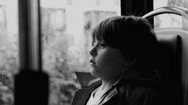 Yağmurlu bir günde otobüsün camından dışarı bakan yağmurluk giymiş bir çocuğun yakın çekimi. Camdaki yağmur damlaları ve bulanık arkaplan sessiz bir tefekkür ve içgözlem havasını vurgular.