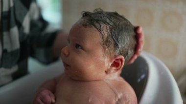 Yeni doğan bir bebeğin kafasının, bebek bakıcısı tarafından nazikçe yıkanırken, bebek yukarı bakarken, güvenli bir ortamda banyo sırasında huzurlu ve hassas bir anı yakalıyor.