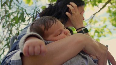 Anne, bebeği huzur içinde uyurken kucağında tutuyor, elini alnında dinlendiriyor, ikisi de ağaçların gölgesinde oturuyordu, sakin bir anı yansıtıyordu.