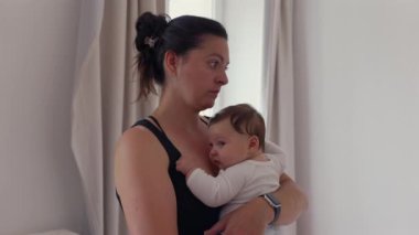 Anne derin bir tefekkür içinde yatak odasında duruyor, bebeği göğsüne yakın tutuyor, sessiz bir bağ ve yansıma anı gösteriyor.