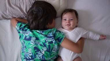 Bebeğin kucağında bebeği tutarken, bebek kameraya bakarken, kardeş yan yana yatarken, iki kardeş arasında şefkatli ve sevgi dolu bir an.