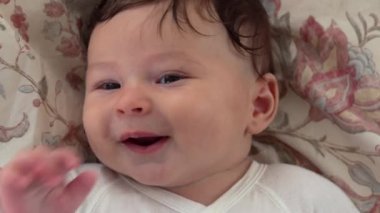 Bebek, çiçekli battaniyeye uzanmış gülümsüyor, bebeğin yüzü rahat ve memnun, bebek, sakin bir ifadeyle usulca bakıyor, bebek huzur içinde dinleniyor ve neşeli görünüyor.