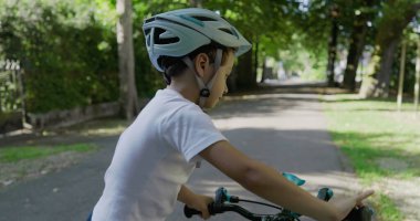 Bisikletiyle parkta gezen, kask takan, önündeki yola odaklanmış, yemyeşil ve büyük ağaçlarla çevrili, açık havada fiziksel aktivitelerle meşgul olan bir çocuk.