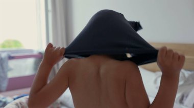 Genç bir çocuk gömleğini başının üstüne kaldırıyor, rahat bir yatak odasında giyiniyor. Günlük hayatın basitliği ve çocukluğun bağımsızlığı.