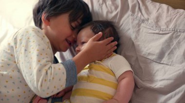 Büyük kardeş yatakta gülümseyen bir bebeği kucaklıyor, sevgi ve bağlılık dolu bir anı paylaşıyor. İçerideki sıcak ortam aile bağlarının sıcaklığını ve kardeş sevgisini yansıtıyor.