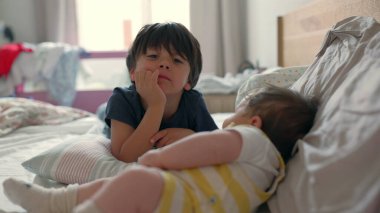 Büyük kardeş yatağın üzerinde bir kardeşin yanında yatıyor, sıcak bir etkileşim içinde. Rahat ortam, kardeşler arasındaki neşeli ve şefkatli bağı yakalar.