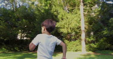 Parkta koşan genç bir çocuk, beyaz bir gömlek giymiş, kararlı bir ifadeyle, yeşilliklerle çevrili, güneşli bir günde dışarıda enerjik bir anın tadını çıkarıyor.