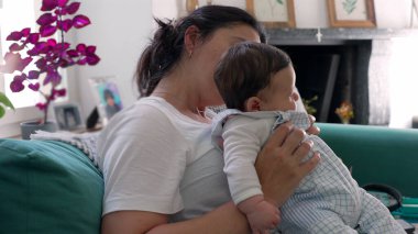 Anne nazikçe bebeğini kucağında tutuyor, her ikisi de ev dekoru ve mor bitkilerle çevrili, sevgi ve birlikteliği simgeleyen, sessiz bir yansımayla aşağıya bakıyor.