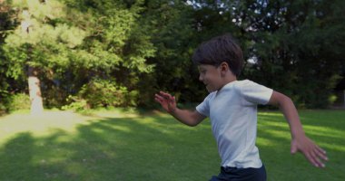 Parkta koşan genç bir çocuk, beyaz bir gömlek giymiş, kararlı bir ifadeyle, yeşilliklerle çevrili, güneşli bir günde dışarıda enerjik bir anın tadını çıkarıyor.