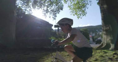 Genç çocuk güneşli bir mahallede bisiklet sürüyor, güvenlik için kask takıyor, azim ve odaklanma gösteriyor.