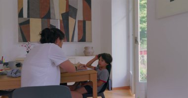 Anne ve oğul ahşap bir masada evde eğitim seansı. Anne, parlak ışıklı bir odada çocuğuna dikkatlice yardım ediyor.