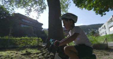 Genç çocuk güneşli bir mahallede bisiklet sürüyor, güvenlik için kask takıyor, azim ve odaklanma gösteriyor.