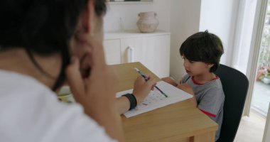 Evde eğitim seansı sırasında masada oturan genç bir çocuk, modern dekorlu iyi ışıklandırılmış bir odada eğitim malzemeleriyle öğretmenlik yapan annesine bakıyor.