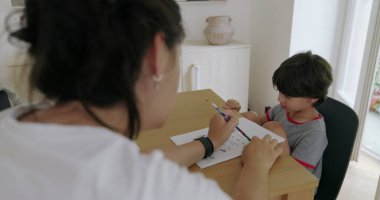 Evde eğitim seansı sırasında masada oturan genç bir çocuk, modern dekorlu iyi ışıklandırılmış bir odada eğitim malzemeleriyle öğretmenlik yapan annesine bakıyor.