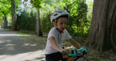 Bisikletiyle parkta gezen, kask takan, önündeki yola odaklanmış, yemyeşil ve büyük ağaçlarla çevrili, açık havada fiziksel aktivitelerle meşgul olan bir çocuk.