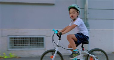 Şehir merkezinde bisiklete binen genç bir çocuğun yan görüntüsü, aracına odaklanmış, kaskla güvenliği vurguluyor ve modern şehir ortamında açık hava aktivitelerinin keyfini çıkarıyor.