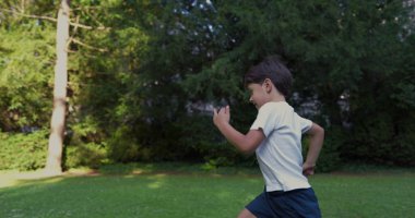 Parkta koşan genç bir çocuk, beyaz bir gömlek giymiş, kararlı bir ifadeyle, yeşilliklerle çevrili, güneşli bir günde dışarıda enerjik bir anın tadını çıkarıyor.