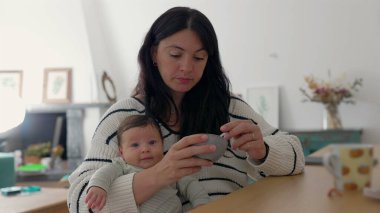 Anne oturma odasında yoğurt yerken bebeğini tutuyor. Samimi ortam ve gündelik an anne ve çocuk arasındaki samimi bağı yansıtıyor.