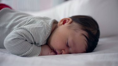 Yeni doğmuş bebek, çizgili tulum giymiş, huzurlu bir şekilde bir yatakta uyuyor. Rahat bir ev ortamında, huzurlu ve huzurlu bir ilk yaşam anı yakalıyor.