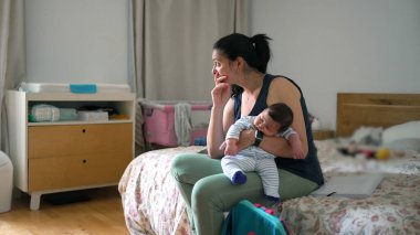 Genç anne uyuyan bebeğini yatakta otururken kucağında tutuyor. Düşünceli, rahat ve samimi bir yatak odası ortamında ona bakıyor.