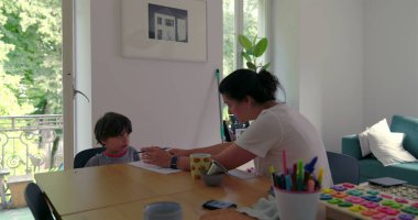 Anne ve oğul, ahşap bir masada, evde eğitim seansı sırasında, anne, doğal ışıkla dolu parlak bir odada öğretirken el kol hareketi yapıyor.