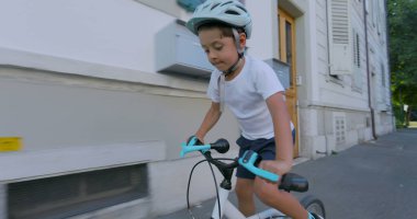 Beyaz kasklı ve beyaz gömlekli, şehir ortamında bisiklet süren genç bir çocuk, önündeki yola odaklanmış. Güneşli bir günün keyfini çıkarırken konut binalarının önünden geçiyor.