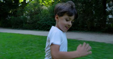 Genç bir çocuk çimenli bir parkta neşeli bir şekilde koşuyor, beyaz bir gömlek giyiyor, mutlu bir ifadeyle, yeşilliklerle çevrili, enerjik ve eğlenceli bir anın tadını çıkarıyor.