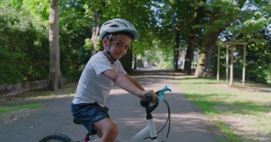 Parkta bisikletli gülümseyen bir çocuk, güvenlik için kask takıyor, ağaçlarla dolu bir yolda duruyor, güneşli bir günün tadını çıkarıyor, gençlik enerjisini sergiliyor.