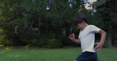 Parkta koşan genç bir çocuk, beyaz bir gömlek giymiş, kararlı bir ifadeyle, yeşilliklerle çevrili, güneşli bir günde dışarıda enerjik bir anın tadını çıkarıyor.