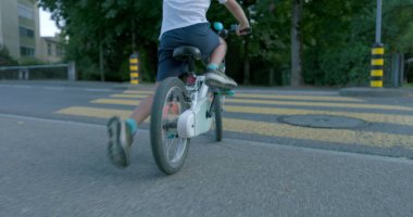 Beyaz bir bisiklete binen, şehir ortamında yaya geçidinden geçen, kasklı ve mavi çoraplı, güvenlik ve aktif yaşam tarzını vurgulayan genç bir çocuğun arka görüntüsü.