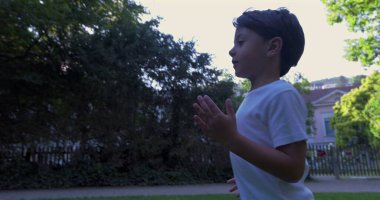 Genç bir çocuk çimenli bir parkta koşuyor, kararlı bir ifadeyle, ağaçlar ve binalarla çevrili, açık havada güneşli bir günün tadını çıkarıyor.