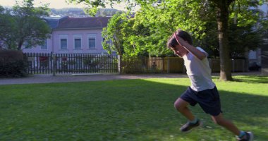 Genç bir çocuk çimenli bir parkta koşuyor, kararlı bir ifadeyle, ağaçlar ve binalarla çevrili, açık havada güneşli bir günün tadını çıkarıyor.