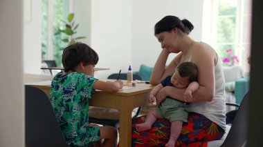 Anne, çocuk resim çizerken ona göz kulak olur, kucağında uyuyan bir bebek, sıcak bir ev ortamında bakım ve şefkat, aile bağı...