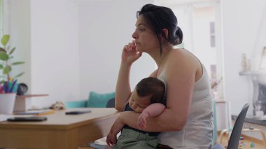 Anne, uyuyan bebeğini kucağında tutarken düşünceli bir şekilde, masada dalgın dalgın bir şekilde, sakin bir ev ortamında anneliğin sessiz ve içsel anlarını yakalıyor.