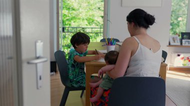 Anne evde eğitim seansı sırasında çocuklarıyla birlikte masada oturur, anne bebeği beşiğinde tutarken gizli eğitim aktiviteleri yapar, huzurlu bir aile anını canlandırır.