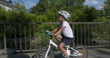 Genç bir çocuk nehir üzerinde bir köprüde bisiklet sürüyor, güvenlik için kask takıyor, arka planda şehir binaları var, azim gösteriyor ve açık hava aktivitelerine odaklanıyor.