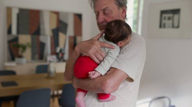 Son sınıf öğrencisi, uyuyan torununu sevgiyle kucağında tutuyor. Huzurlu bir yuva ortamında bebeği nazikçe kucaklıyor. Aile bağının ve nesiller arası bağın iç açıcı bir sahnesi.